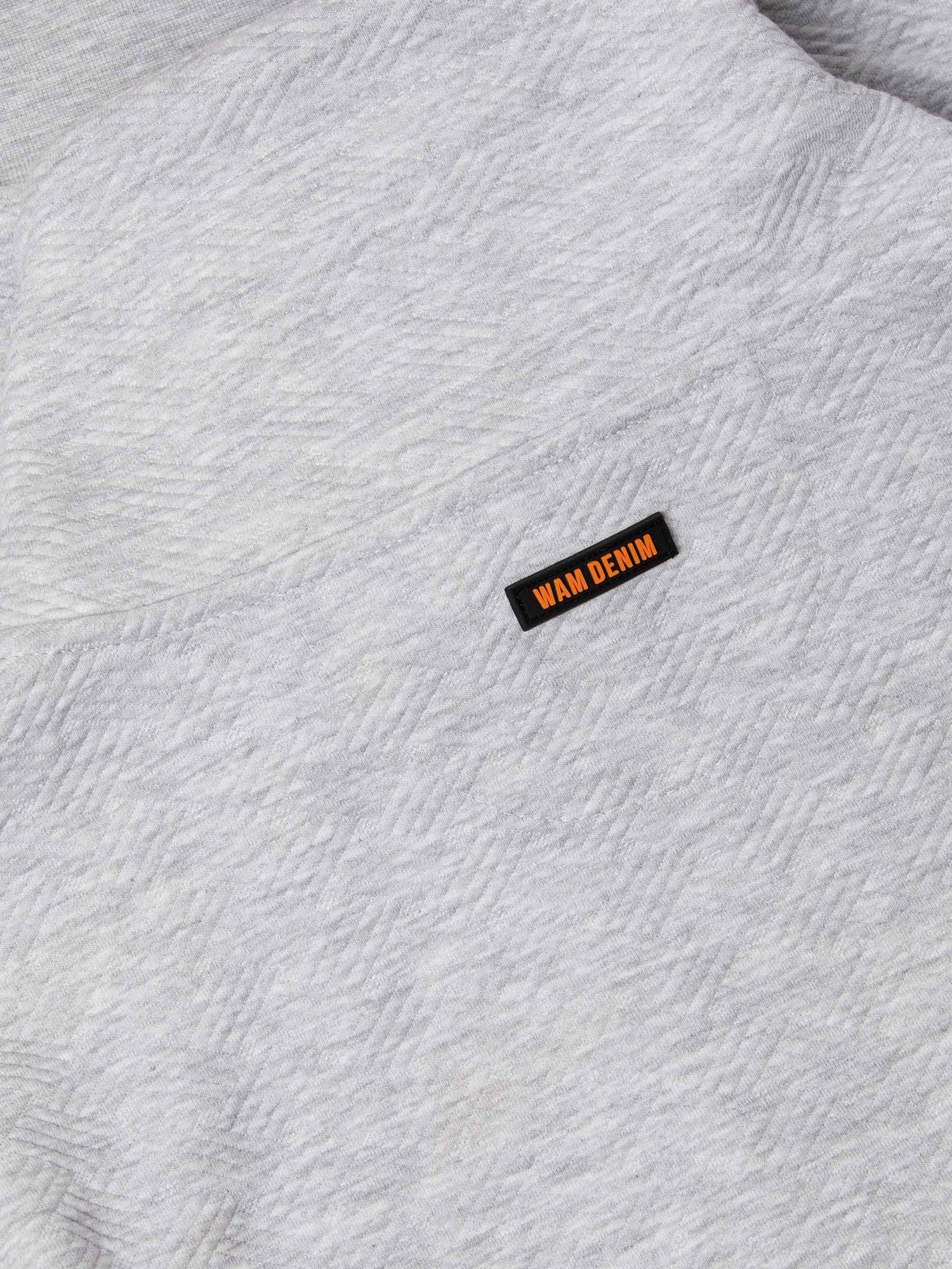 Close-up of light grey textured fabric featuring black and orange Wam Denim logo tag – Wam Denim.